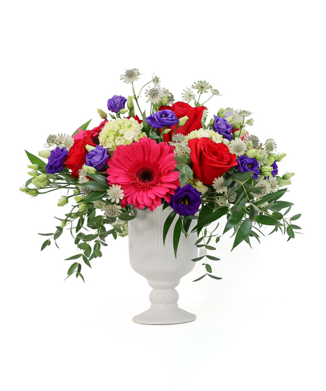 A bright mix of pink gerbera daisies, red roses, and purple lisianthus, with lime-green hydrangea and white astrantia designed in a white ceramic vase..