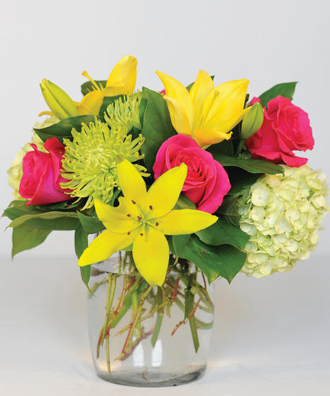 An arrangement of yellow lilies, hot pink roses, green spider mums, white hydrangeas and green foliage in a clear glass vase.
