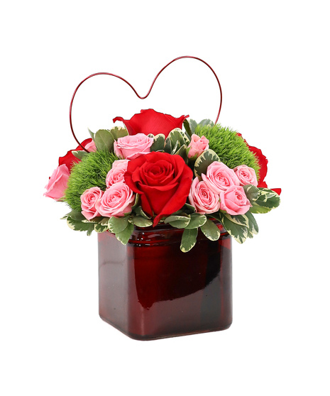 A mounded floral arrangement featuring red roses, pink spray roses, green trick, and fresh foliage, designed in a red cube vase with a wire heart accent.