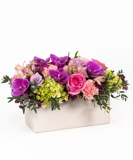 A white rectangular container with purple orchids, lavender roses, pink carnations, pink alstroemeria, and green hydrangea.