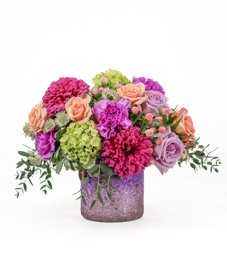 A compact, colorful floral arrangement in a lavender container featuring peach roses, pink and purple blooms, green hydrangea, and soft greenery.
