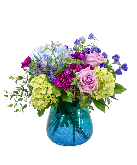 Blue and green hydrangeas, lavender roses, purple carnations, and greenery in a blue glass container.