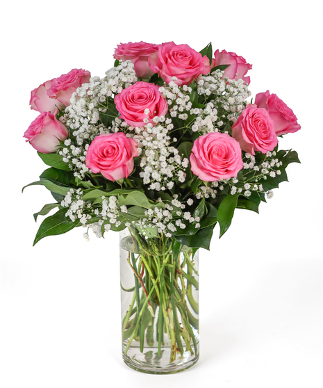A clear vase filled with twelve pink roses, accented with white baby’s breath and green foliage.