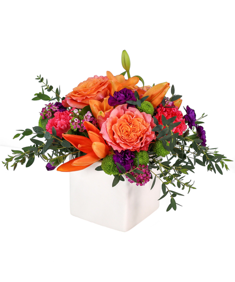 Bright mixed flower arrangement in a white cube with orange roses, pink carnations, green button mums, purple mini carnations, and waxflower.