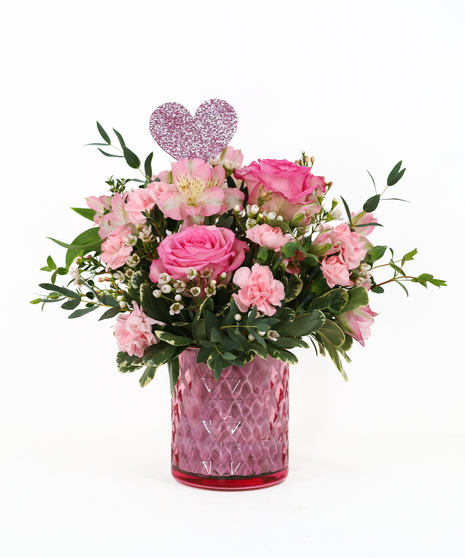 A rounded pink floral arrangement featuring roses, soft blush blooms, fresh greenery, and a pink glass vase with a subtle heart accent.