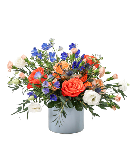Orange and peach roses with blue delphinium, white lisianthus, peach mini carnations, greenery, and monarch butterflies in a soft blue cylinder vase.