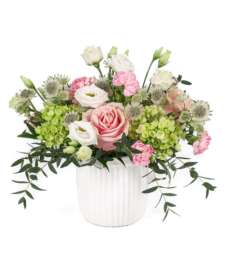 A white container filled with a soft pink rose, pink and white flowers, green hydrangea, and flowing greenery.