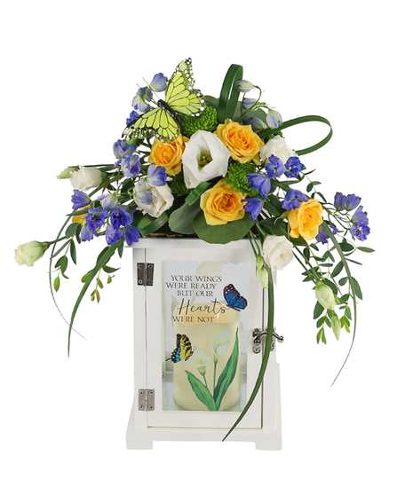 Sympathy floral arrangement with yellow roses, blue accent flowers, and a butterfly topper on a white memorial lantern with trailing greenery.