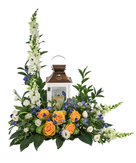 Sympathy floral arrangement with tall white snapdragons, yellow roses, and blue accent flowers surrounding a white memorial lantern with a bronze top, set in lush greenery.