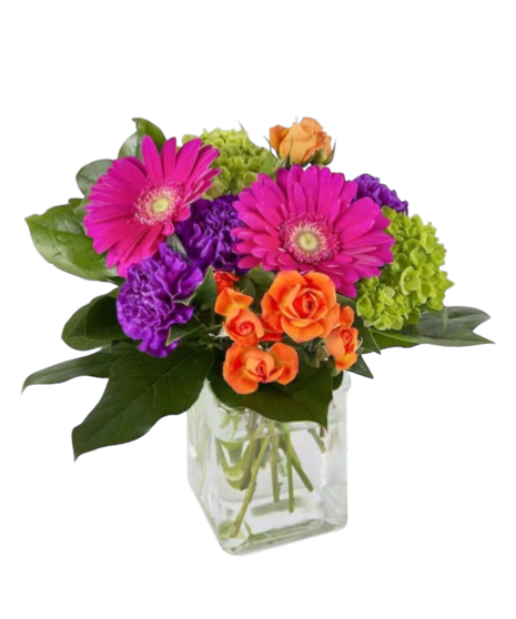 A mix of hot pink gerbera daisies, purple carnations, and small orange roses with green hydrangea and foliage arranged in a clear glass cube container