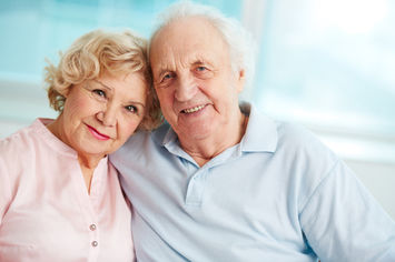 An elderly couple, seated closely together and smiling warmly, embraces in a bright, airy room with large windows in the background.